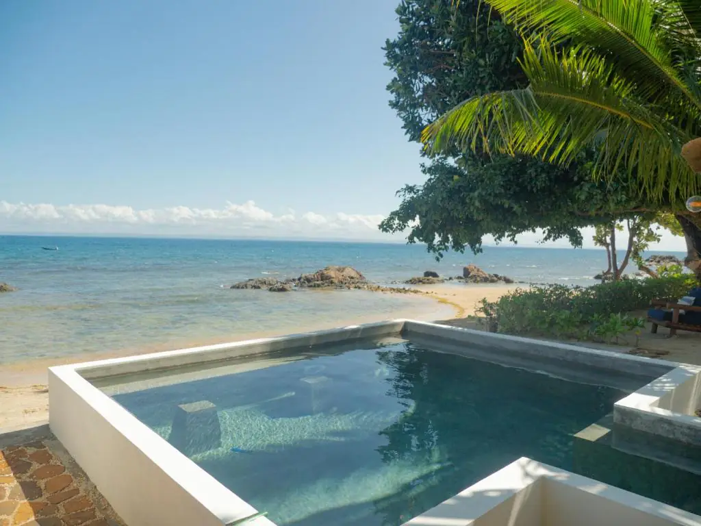 La piscine blanche installée sur la plage, l'eau du bassin dans le prolongement du lagon et des rochers affleurants.