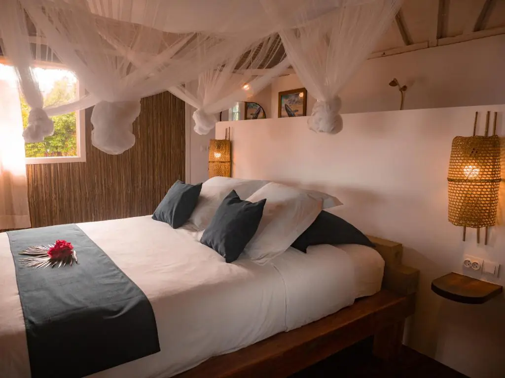 Détail du lit : linge blanc, coussins bleu nuit, chemin de lit ardoise et une fleur d'hibiscus rouge posée dessus.