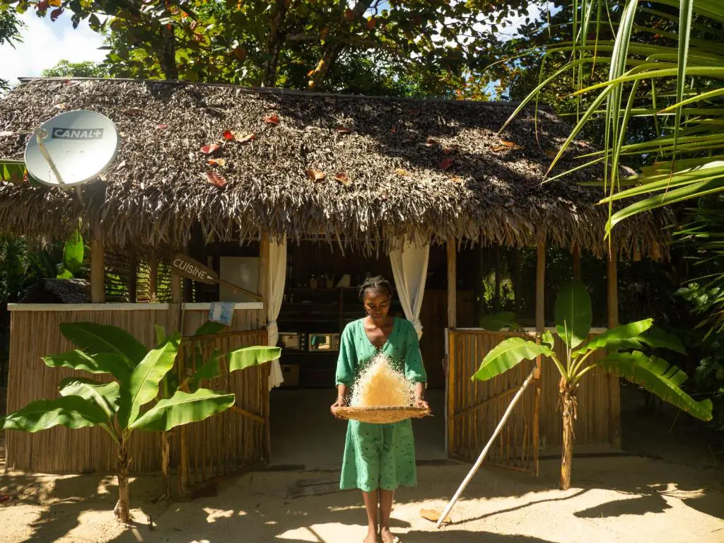Devant la cuisine au toit de palme, une membre de l'équipe vanne du riz dans un plateau tressé traditionnel.