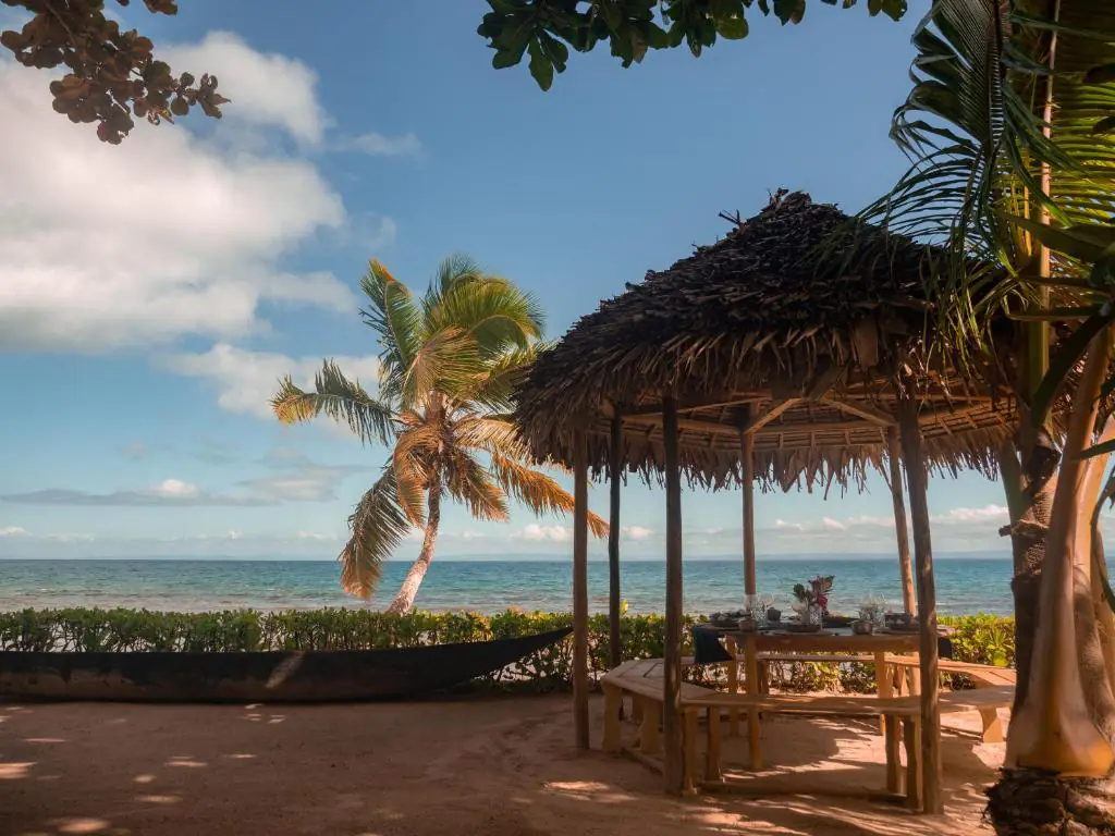 La paillote au toit de palme posée sur le sable, une pirogue à côté et deux cocotiers penchés vers le lagon.