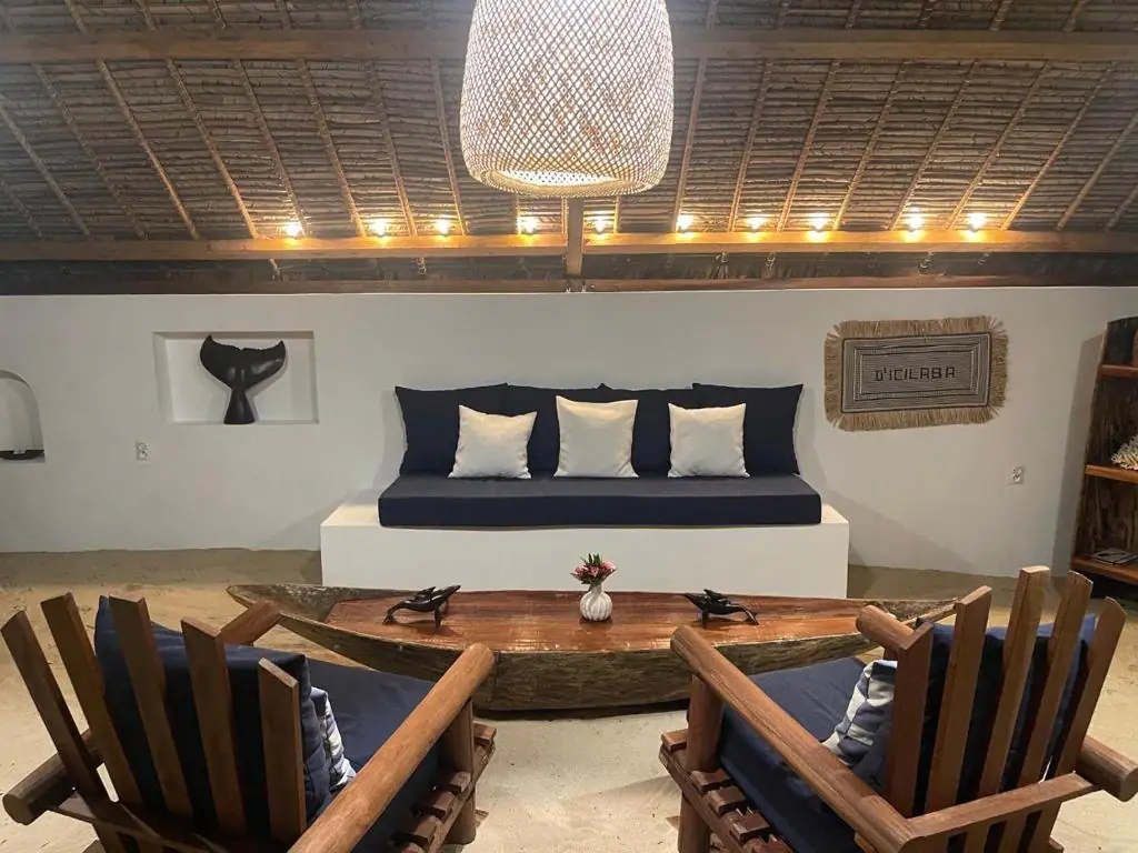 Le salon de plage : banquette bleu nuit à coussins blancs, pirogue en bois servant de table basse, grand luminaire tressé et queue de baleine sculptée dans une niche murale.
