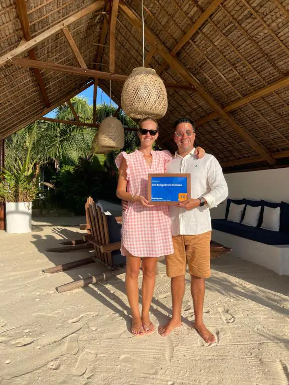 Emilie et Floryan, les propriétaires, pieds nus sur le sable sous le salon de plage, tenant le trophée Traveller Review Awards de Booking.com.