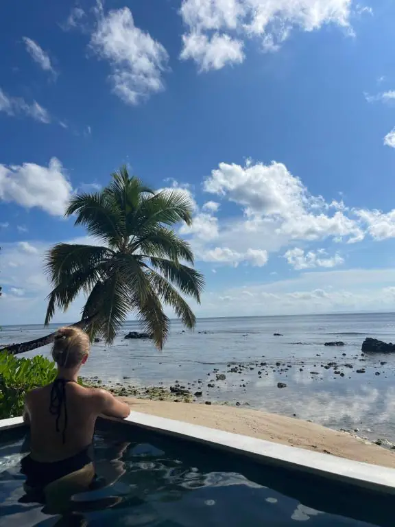 Une personne accoudée au rebord de la piscine, regardant l'océan et un cocotier penché au-dessus du sable.