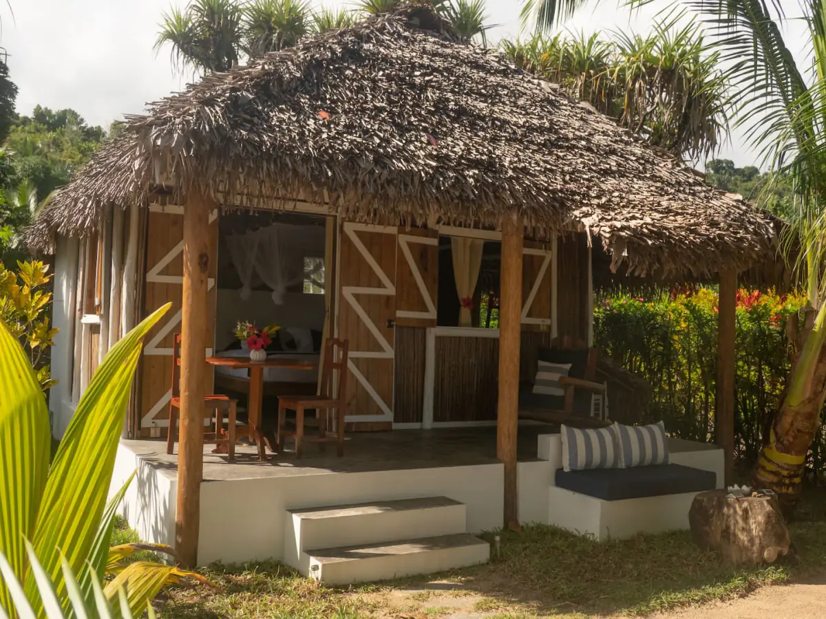 Le bungalow vu du jardin : façade en bois à motifs zigzag blancs, toit de palme, terrasse avec table et banquette d'extérieur.