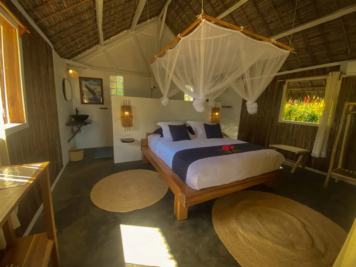 Intérieur du bungalow : grand lit en bois massif sous une moustiquaire à baldaquin, murs de bambou tressé et charpente en palme apparente.