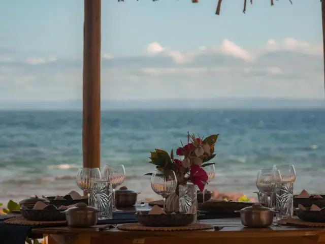 Table dressée sous la paillote, verres en cristal et fleurs, l'océan flouté en arrière-plan.