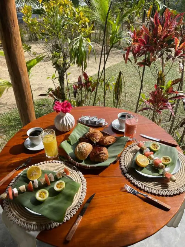 Petit-déjeuner vu de dessus : assiettes sur feuilles de bananier, brochettes de fruits, viennoiseries et café.