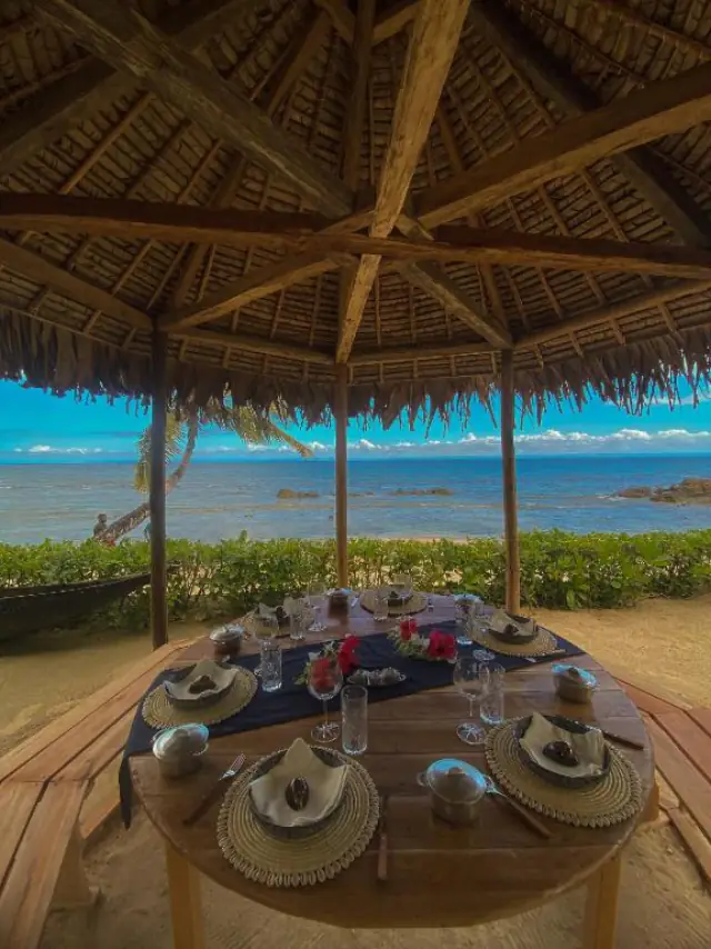 Vue depuis l'intérieur de la paillote : la table dressée, encadrée par les poteaux en bois, face au lagon et à la pirogue échouée.