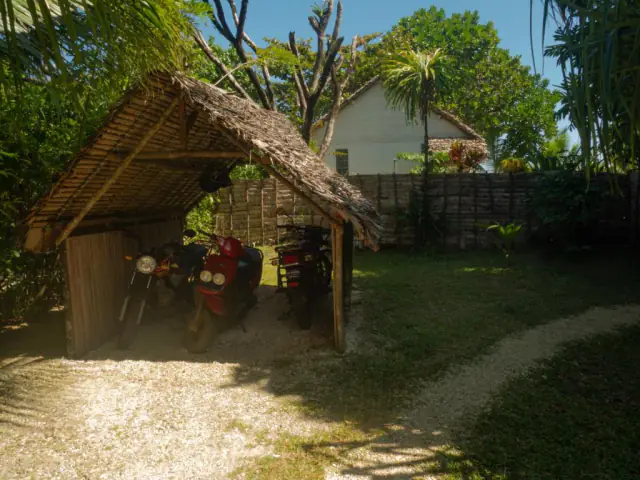 Abri en bois au toit de palme abritant deux motos et un scooter, dans le jardin de la propriété.
