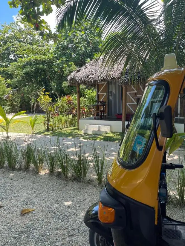 Un tuk-tuk jaune stationné devant le bungalow, sur l'allée de gravier bordée de plantes.
