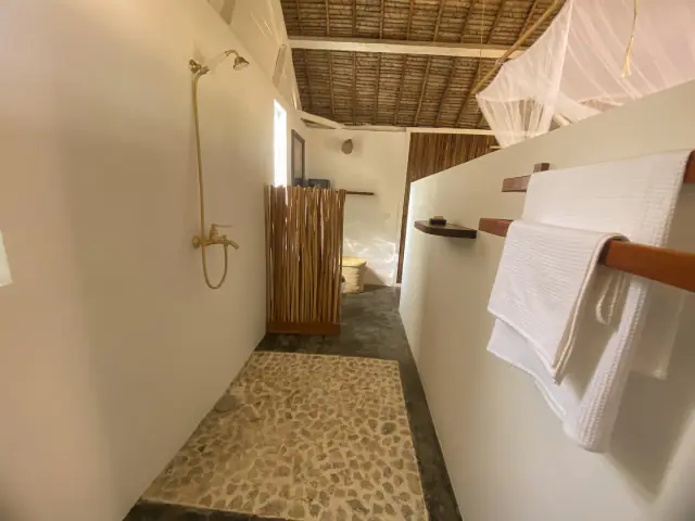 La salle d'eau vue dans sa longueur : douche laiton, paravent de bambou et serviettes blanches sur une barre en bois.