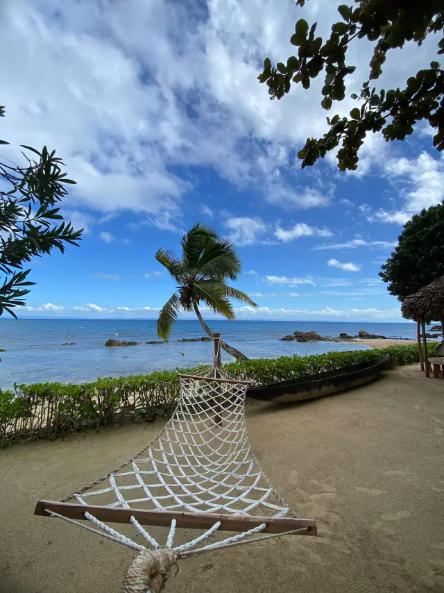 Un hamac blanc tendu sur le sable, une pirogue noire et un cocotier penché au-dessus du lagon bleu.