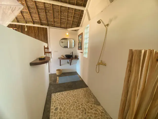 La salle d'eau ouverte : vasque noire, miroir arrondi, paravent en rondins de bois et lumière naturelle par une brique de verre.